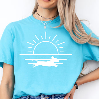 Lagoon blue shirt with a graphic of a dachshund running in front of the ocean at sunset.