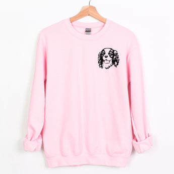 Light Pink Cavalier King Charles Spaniel sweatshirt on a wood clothes hanger. Sweatshirt features a Cavalier face printed on the left chest in black.