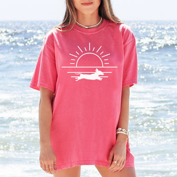 Pink dachshund summer shirt with a graphic of a dachshund running in front of the ocean and a sunset modeled by a woman standing on the beach in front of the ocean.
